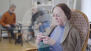 Close-up portrait of mature Caucasian woman examining knitting. Old female retiree practicing hobby in nursing home.