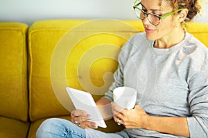 Close up portrait of mature pretty woman reading a book on tablet with glasses sitting on the yellow couch at home in break