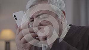 Close-up portrait of mope-eyed senior man swiping smartphone screen. Old Caucasian retiree using phone indoors. Modern