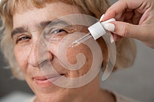 Close-up portrait of an old woman applying hyaluronic acid serum with a pipette. Anti-aging face care.