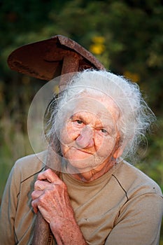 Close-up portrait of an old woman with gray hair smiling and looking at the camera, holding a rusty shovel in her hands, face in