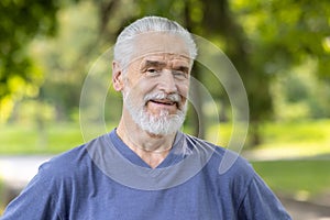 Close-up portrait of an older gray-haired man standing in a park in sportswear and smiling at the camera