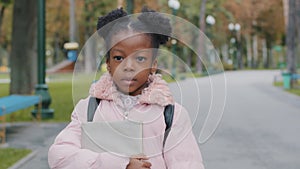 Close-up portrait outdoors serious sad little cute african american schoolgirl holding notebook textbook book walking to