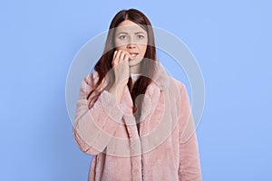 Close up portrait of pensive worried scared young woman biting her nails isolated over blue background, female wearing pale pink