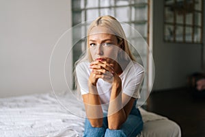 Close-up portrait of pensive young blonde woman sitting alone in living room and sad looking at camera holding hands on