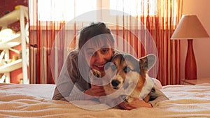 Close-up portrait of pleased woman lying on bed at home with her loved dog Welsh Corgi.