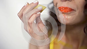 Close up portrait of plump wet lips with bright orange lipstick biting sour piece of lemon. Slow motion