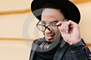 Close-up portrait of positive african man playfully touching his glasses. Outdoor shot of elegant black male model in