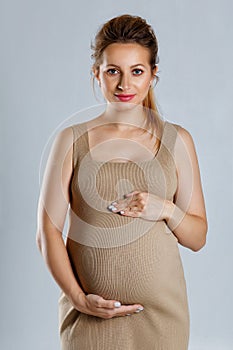 Close-up portrait of a pregnant woman in a beige dress hugging her stomach with both hands on a gray background and looking at it