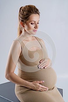 Close-up portrait of a pregnant woman in a beige dress hugging her stomach with both hands on a gray background and looking at it