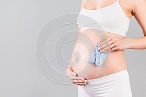 Close up portrait of pregnant woman in white underwear holding blue socks for a baby boy at gray background. Future child