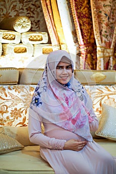 Close up portrait of pregnant young Asian muslim woman wearing colorful hijab sitting on coach with golden backdrop sheet