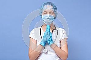 Close up portrait of pretty female with her hands together and praying before patient, keeing eyes closed, wearing medical