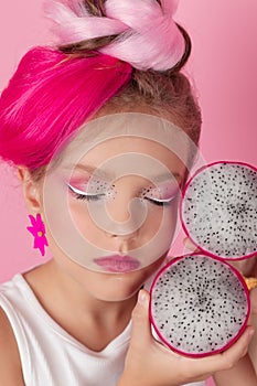 Close-up portrait of pretty girl with pink hairstyle with dragon fruit on pink background. Studio shot of charming tween
