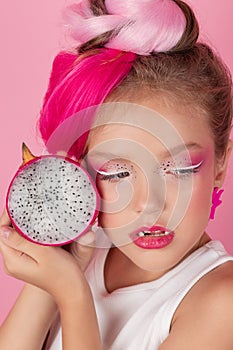 Close-up portrait of pretty girl with pink hairstyle with dragon fruit on pink background. Studio shot of charming tween