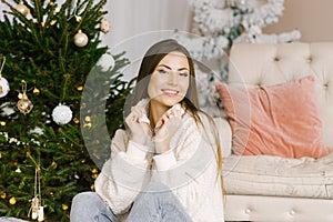 Close-up portrait of a pretty girl sitting near a Christmas tree and lights. She is happy and smiling in the cozy living room of