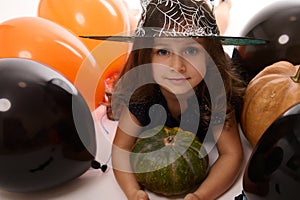 Close-up portrait of pretty little girl in carnival witch costume and wizard hat looking at camera posing with pumpkin in the