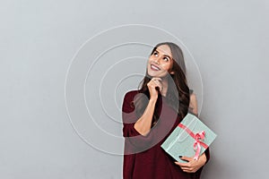 Close-up portrait of pretty smiling woman in stylish knitted sweater touching her chin while holding gift box, looking aside