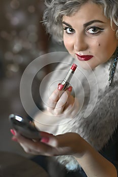 Close up portrait of pretty woman correcting makeup painting lips with red lipstick holding mirror in her hand. Retro