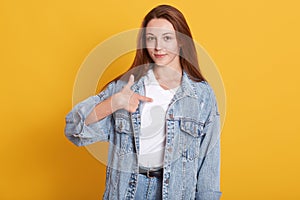 Close up portrait of pretty young woman with long dark staright hair wearing denim jacket and white shirt, standing and pointing