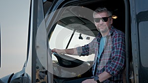 Close up Portrait of Professional Truck Driver sits in his truck and Behind Him Parked Long Haul Semi-Truck.