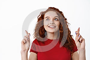 Close up portrait of redhead hopeful woman with curly hair, cross fingers for good luck, staring at upper left corner
