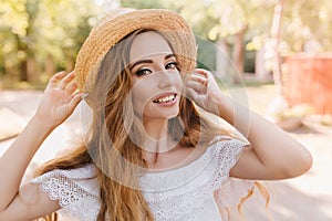 Close-up portrait of refined lady in trendy summer hat smiling on blur nature background. Outdoor photo of pretty long