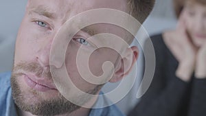 Close up portrait of sad unhappy young man looking away in the foreground. The blurred figure of mature lady holding her