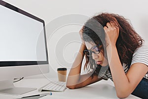 Close-up portrait of sad young woman lies near computer with white screen and cup of coffee. Indoor photo of tired girl