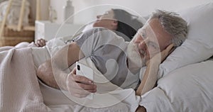 Close-up portrait of senior Caucasian man lying in bed and using smartphone as his wife sleeping at the background