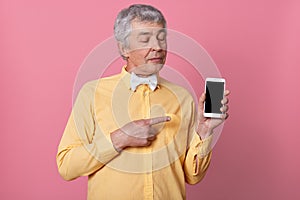 Close up portrait of senior man wearing yellow shirt with white bowtie showing black blank smart phone screen, looking at his