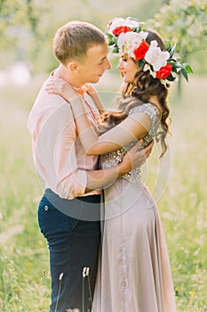 Close-up portrait of sensual blonde woman with flowers wreath and handsome man wearing peach shirt hugging face-to-face in the ga