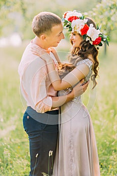 Close-up portrait of sensual blonde woman with flowers wreath and handsome man wearing peach shirt hugging face-to-face in the ga