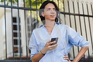 Close-up portrait of serious attractive woman with mobile phone looking away and waiting friend on the city street. Young brunette
