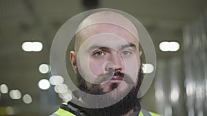 Close-up portrait of serious baldheaded caucasian man with black beard looking at camera. Employee in green vest