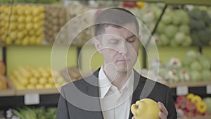 Close-up portrait of serious Caucasian man choosing pear in grocery shop. Handsome guy smelling yellow fruit with