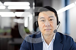 Close-up portrait of a serious young Asian man wearing a headset sitting in the office and looking confidently into the