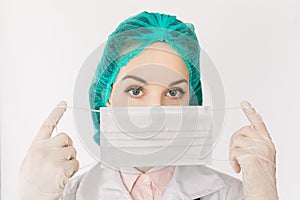 Close-up portrait of serious young nurse or doctor in cap, gloves and white coat, showing how to wear medical face mask