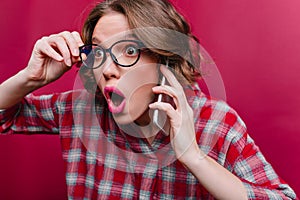 Close-up portrait of shocked girl in glasses talking on phone. Short-haired young woman in checkered shirt posing on