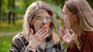 Close-up portrait of shocked surprised Caucasian woman in sunny autumn park with friend appearing whispering rumors on