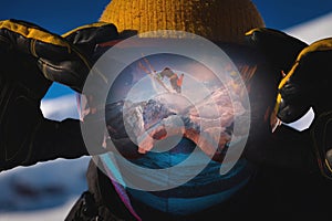 Close-up portrait of a skier in a mask holding on to ski goggles in the reflection of which the skier does a backflip