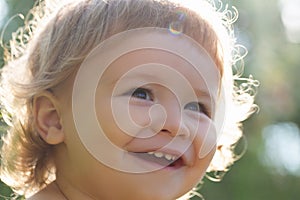Close up portrait of a small blond baby boy. Funny kids cropped face outdoor on sunny day. Kids smiling, cute smile.