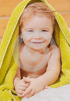 Close up portrait of a small blond boy cover body under towel after bath. Funny baby after bathing on bed at home.