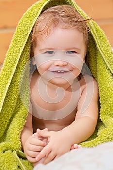 Close up portrait of a small blond boy cover body under towel after bath. Funny baby after bathing on bed at home.