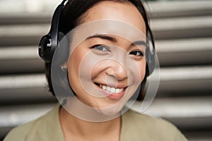 Close up portrait of smiling asian girl in headphones, listens to music outdoors, looking happy.