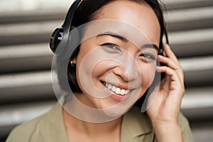 Close up portrait of smiling asian girl in headphones, listens to music outdoors, looking happy.