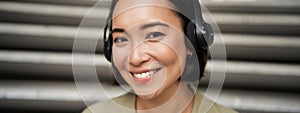 Close up portrait of smiling asian girl in headphones, listens to music outdoors, looking happy.