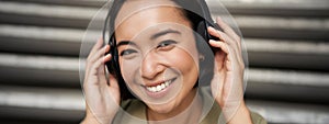 Close up portrait of smiling asian girl in headphones, listens to music outdoors, looking happy.