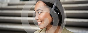 Close up portrait of smiling asian girl in headphones, listens to music outdoors, looking happy.