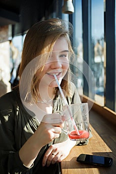 Close up portrait of smiling beautiful teen student girl with a glass mug drinks through a straw fruit tea at street cafe